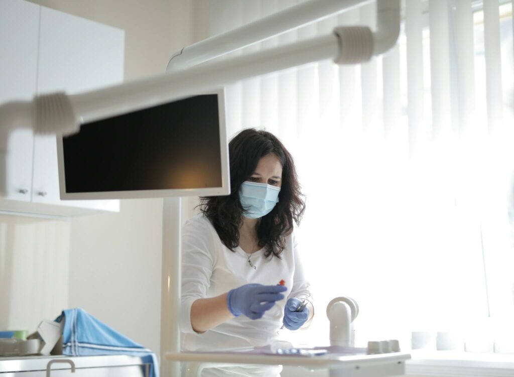 Serious female dentist in medical mask and latex gloves wearing white uniform preparing instruments for treatment in light modern dental room in clinic